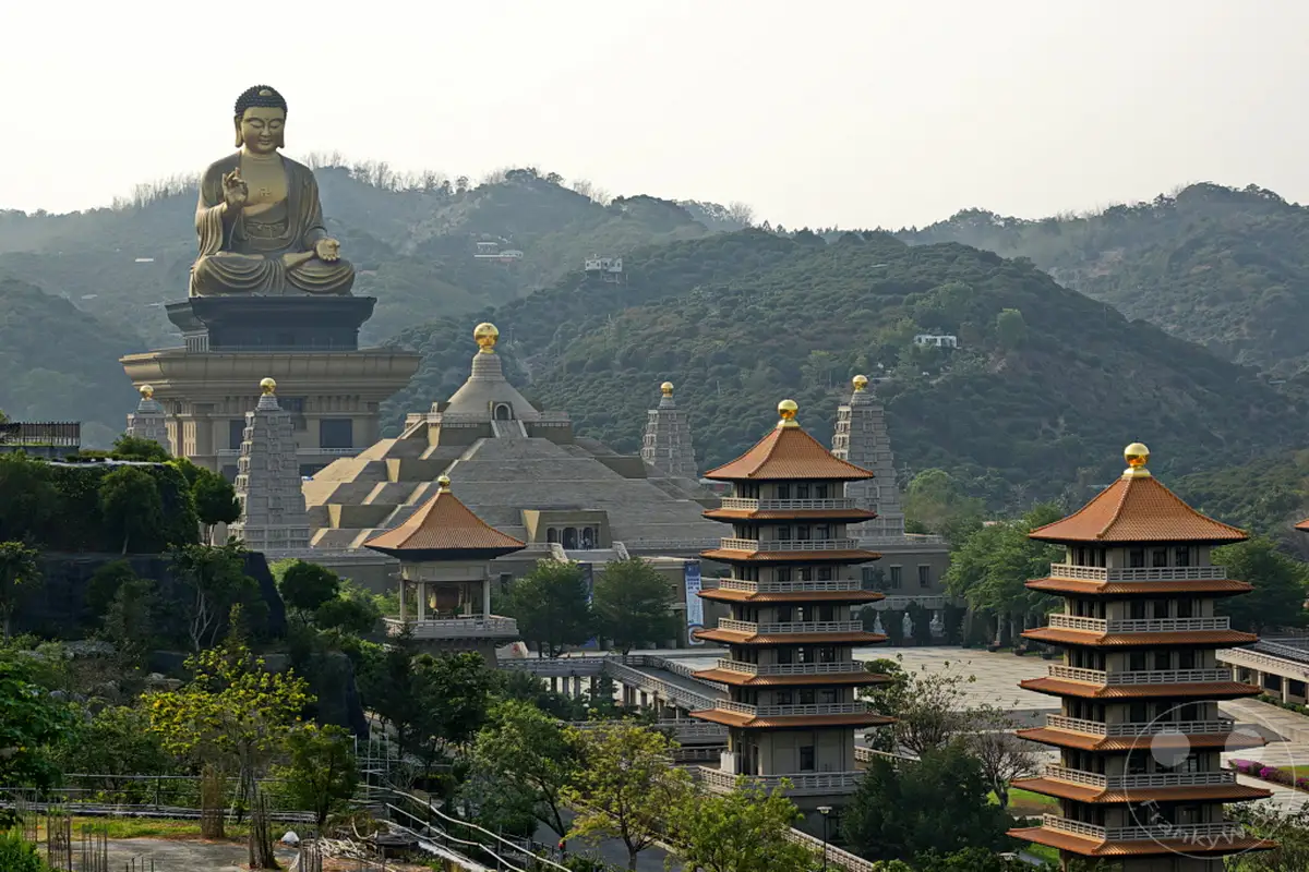 Die monumentale Buddha-Statue thront über einem weitläufigen Tempelkomplex in Taiwan. Mehrere mehrstöckige Pagoden mit orangefarbenen Dächern und goldenen Spitzen sind im Vordergrund und Mittelgrund zu sehen. Die Szenerie strahlt eine tiefe Ruhe und Erhabenheit aus, eingebettet in die sanften Hügel des Landes.