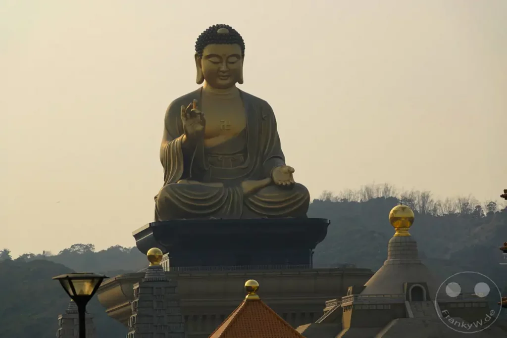 Die monumentale Buddha-Statue in Fo Guang Shan, Kaohsiung, Taiwan, strahlt in goldenem Glanz gegen einen pastellfarbenen Himmel. Umgeben von sanften Hügeln und architektonischen Elementen des Buddha Museums, vermittelt die Szene eine Atmosphäre der Ruhe und Erhabenheit. Die Statue sitzt in meditativer Haltung auf einem Sockel, während im Vordergrund eine Laterne und weitere Gebäude des Komplexes sichtbar sind.