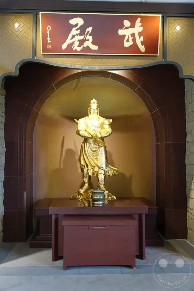 Im Fo Guang Shan Buddha Museum in Kaohsiung, Taiwan, steht eine imposante goldene Buddha-Statue im Mittelpunkt einer Nische. Die Statue strahlt eine friedliche und ehrwürdige Atmosphäre aus und wird von kunstvollen architektonischen Elementen eingerahmt. Eine dunkle Holztruhe steht davor und vervollständigt die Szene des stillen Ortes.