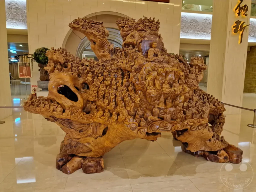 Eine monumentale Holzskulptur mit unzähligen kleinen Buddha-Figuren, die sich über die gesamte Oberfläche schlängelt, ist das Herzstück dieser Aufnahmemöglichkeit im Fo Guang Shan Buddha Museum in Kaohsiung, Taiwan. Die filigrane Handwerkskunst, welche die gesamte Struktur bedeckt, erzeugt eine Atmosphäre der Ruhe und Verehrung, während das natürliche Holz eine erdige Wärme ausstrahlt. Die Szene vermittelt den Eindruck von tausenden Gläubigen, die sich um eine zentrale Buddha-Figur versammeln und so die spirituelle Bedeutung des Ortes unterstreichen.