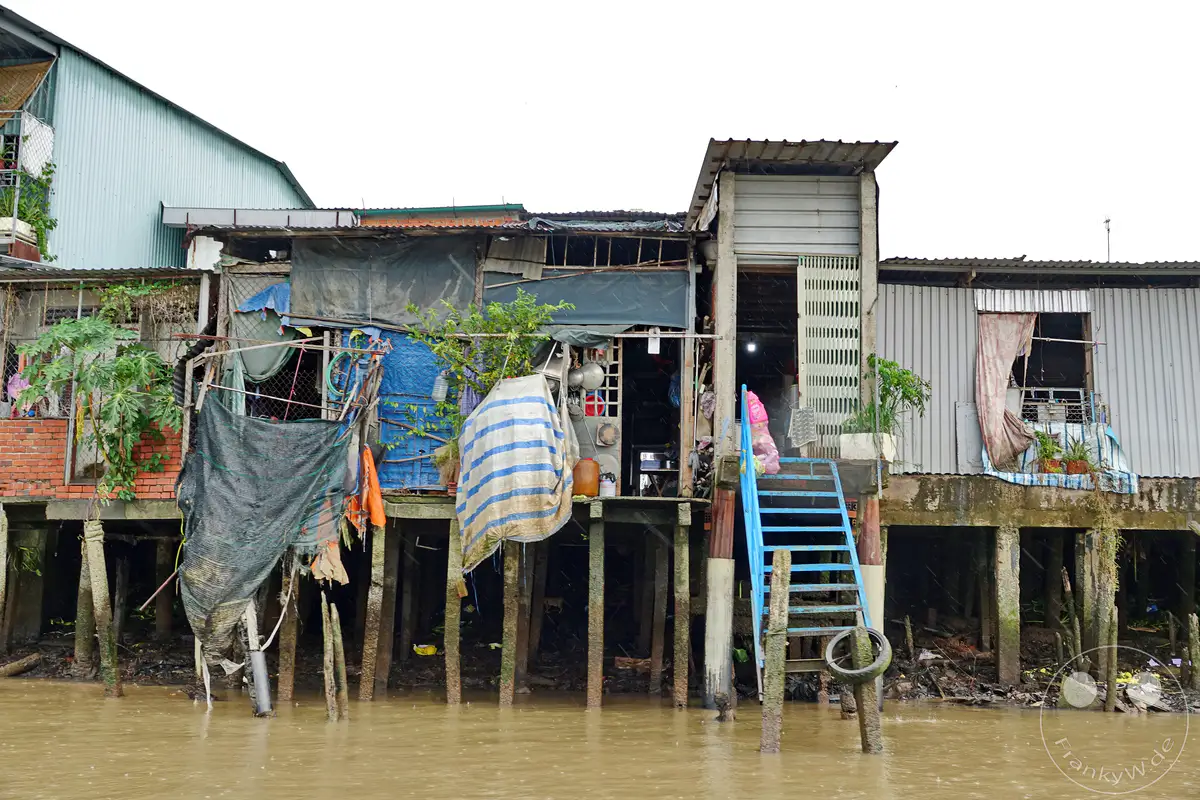Ein provisorisches Stelzenhaus-Dorf am Ufer des Mekong Deltas in Vietnam. Die baufälligen Unterkünfte spiegeln die Armut der Bewohner wider, während das trübe Wasser und der Regen eine melancholische Atmosphäre erzeugen. Dieser Anblick vermittelt einen Eindruck vom rauen Leben am Fluss.