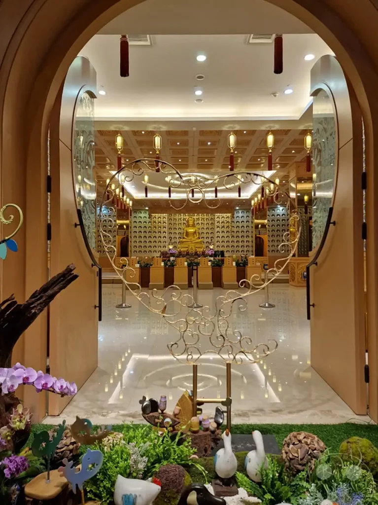 Ein Blick durch einen kunstvoll verzierten Eingang auf den Fo Guang Shan Buddha Museum in Kaohsiung, Taiwan. Die strahlende Buddha-Statue in der Mitte des Tempels wird von einer Wand mit zahlreichen kleineren Buddha-Figuren eingerahmt, während die aufwendige Deckenbeleuchtung und die Dekoration eine würdevolle und friedliche Atmosphäre schaffen. Vor dem Eingangsbereich bietet eine detailreiche Gartendekoration mit Figuren und Blumen einen farbenfrohen Kontrast zur spirituellen Stätte.
