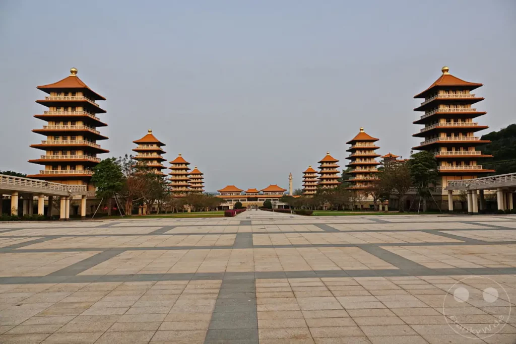 Das Fo Guang Shan Buddha Museum in Kaohsiung, Taiwan, präsentiert sich als symmetrisches architektonisches Meisterwerk mit mehreren Pagoden, die einen zentralen Palast flankieren. Die weitläufige, gepflasterte Vorfläche und der klare Himmel erzeugen eine Atmosphäre der Ruhe und Erhabenheit inmitten der beeindruckenden buddhistischen Anlage. Die symmetrische Anordnung der Gebäude und die friedliche Stimmung laden zur Besinnung ein.