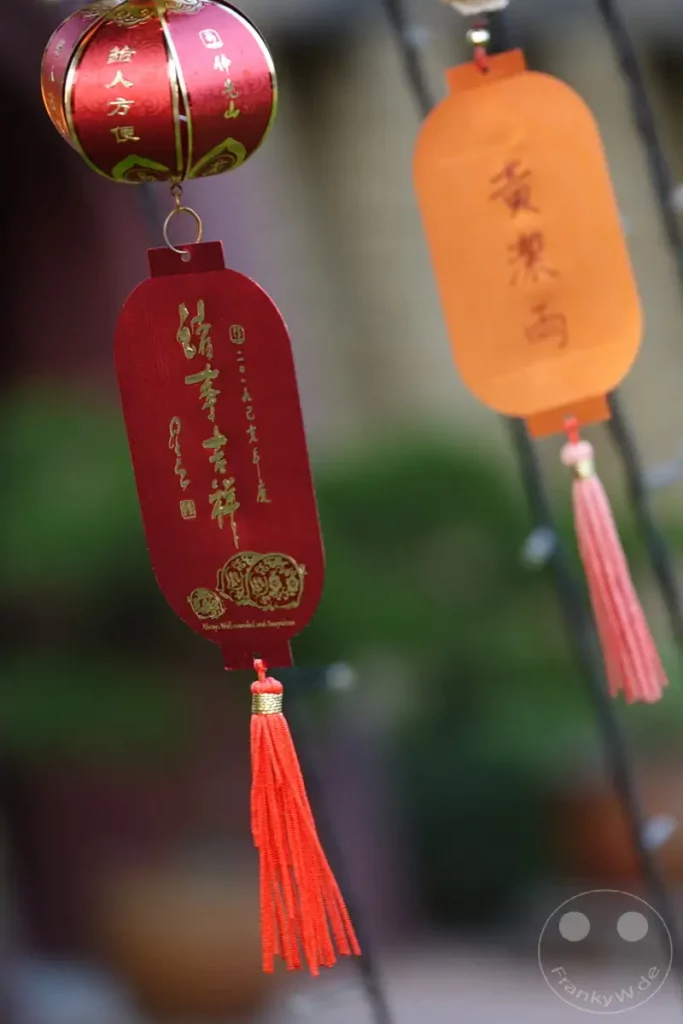 In Kaohsiung, Taiwan, hängen rote und orangefarbene Lampions vor dem Fo Guang Shan Buddha Museum, die mit traditionellen Schriftzeichen und Glückssymbolen verziert sind. Rote Quasten hängen von den Lampions und vermitteln eine festliche Atmosphäre. Das Bild fängt die kulturelle Ästhetik der Dekorationen ein und lässt auf ein geschäftiges Treiben schließen, vielleicht während eines Festes oder einer besonderen Veranstaltung.
