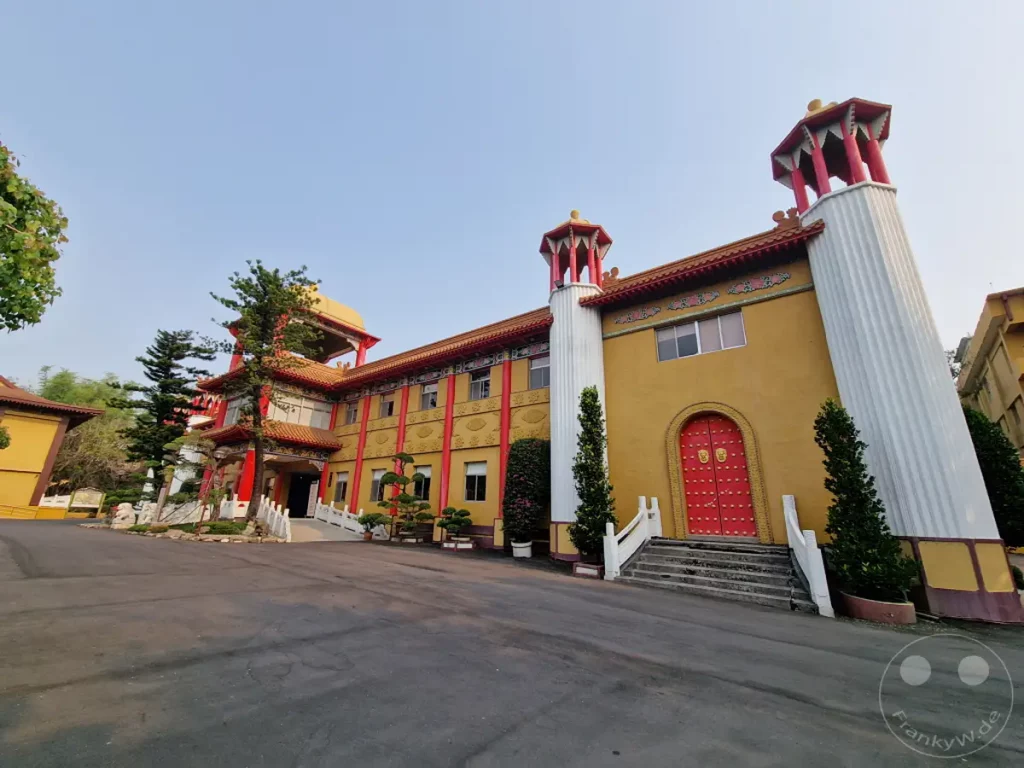 Das Fo Guang Shan Monastery in Kaohsiung, Taiwan, präsentiert sich in leuchtendem Gelb mit auffälligen roten Türen und traditionellen roten Zierleisten. Majestätische weiße Säulen flankieren den Eingang, während Bäume und gepflegte Pflanzen eine friedliche Atmosphäre umgeben. Die Architektur vereint traditionelle Elemente mit monumentaler Gestaltung vor einem klaren Himmel.