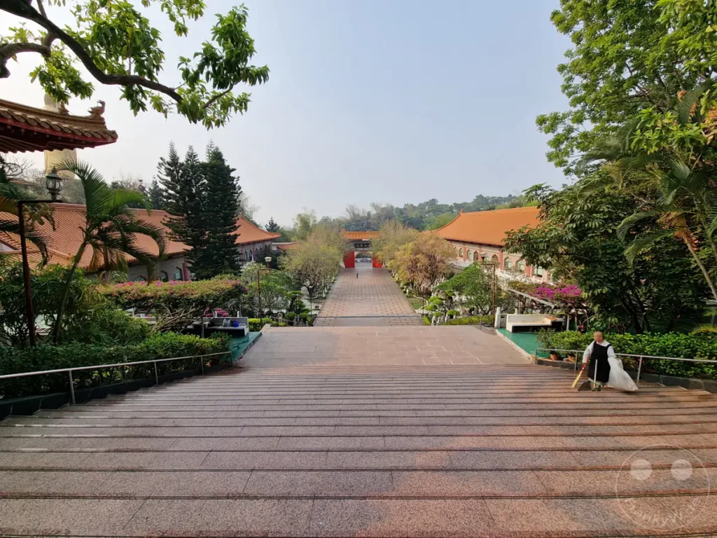 Eine weitläufige Treppenanlage führt zu den Gebäuden des Fo Guang Shan Monastery in Kaohsiung, Taiwan. Eine Person in traditioneller Robe ist mit einem Besen und einem Müllsack beschäftigt, die Umgebung zu säubern. Die Szenerie strahlt eine Atmosphäre der Ruhe und Hingabe aus, umgeben von üppiger Vegetation und traditioneller Architektur.