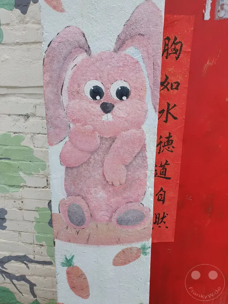 Eine fröhliche rosa Bunny-Wandmalerei ziert eine Säule in Tainan, Taiwan. Die bunte Illustration zeigt das Kaninchen sitzend auf einer Karotte, umgeben von weiteren Gemüsen und einem traditionellen roten Schriftzug. Die verspielte Darstellung vermittelt eine unbeschwerte und festliche Atmosphäre, passend zum chinesischen Neujahrsfest oder Ostern.