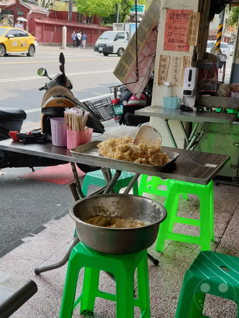Eine Essensauslage an einer Straße in Tainan, Taiwan, bietet Streetfood an. Mehrere grüne Plastikhocker und eine Metallschüssel mit Nahrungsmitteln dominieren die Szene. Die Atmosphäre ist die eines typischen Imbissstandes im Freien.