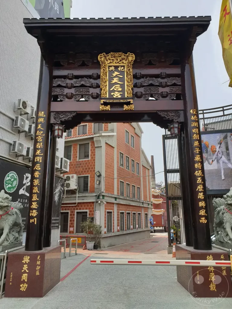 Ein aufwendig gestaltetes traditionelles Tor aus dunklem Holz steht im Vordergrund und gibt den Blick auf eine Straße in Tainan, Taiwan, frei. An den Seiten des Tores sind goldene Schriftzeichen angebracht, die auf die Bedeutung des Ortes hinweisen. Ziegelsteingebäude und eine Tempelarchitektur sind im Hintergrund zu erkennen, die eine Mischung aus Moderne und Tradition vermitteln.