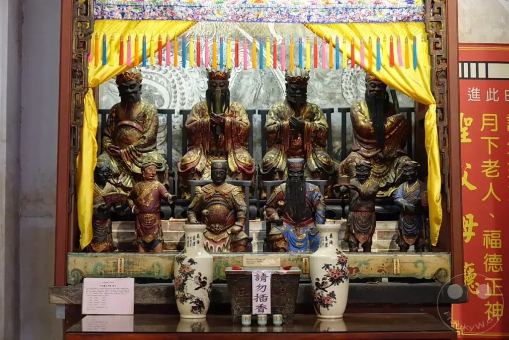 Im Inneren des Tainan Grand Mazu Tempels in Taiwan enthüllt ein reich verzierter Altar eine Reihe von detailreichen Statuen, die Götter und historische Figuren darstellen. Die Figuren sitzen auf Podesten, umgeben von Gelb und farbenfrohen Quasten, was eine festliche und ehrfurchtsvolle Atmosphäre schafft. Der Schrein ist mit traditionellen Keramikvasen und einem Weihrauchfass geschmückt und unterstreicht die spirituelle Bedeutung des Ortes.