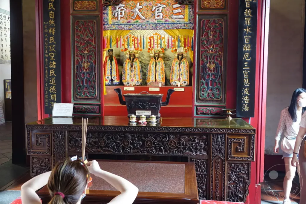 Im Inneren des Tainan Grand Mazu Tempels in Taiwan stehen vier Götterfiguren, geschmückt mit prächtigen Gewändern und Kopfbedeckungen, auf einem Altar. Vor ihnen befindet sich ein großer Räucherkessel, flankiert von kleineren Opfergaben, während im Vordergrund eine Person mit Räucherstäbchen betet. Die Szenerie strahlt eine Atmosphäre tiefer Spiritualität und kultureller Tradition aus.