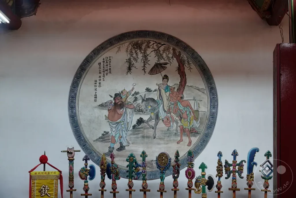 In der Nähe des Tainan Grand Mazu Tempels in Taiwan hängt eine kreisrunde Tempelmalerei, die eine Szene mit mehreren Figuren und einem Esel darstellt. Die Malerei ist mit chinesischen Schriftzeichen verziert und wirkt in ihrer Ausführung traditionell. Unterhalb der Malerei sind mehrere kunstvoll gestaltete, fahnenähnliche Objekte ausgestellt, die ebenfalls religiöse Bedeutung haben könnten.