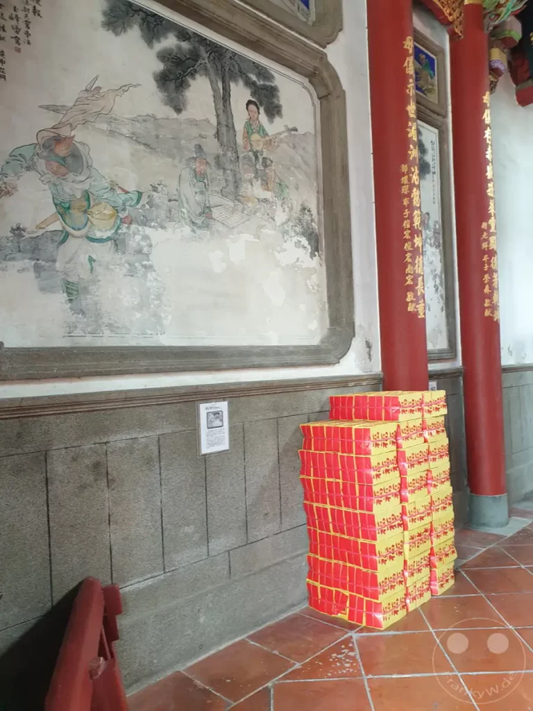 Im Tainan Grand Mazu Temple in Taiwan stapelt sich auf dem Boden ein Berg aus roten und gelben Opfergeld-Stapeln. An der Wand hinter dem Opfergeld ist eine detailreiche Tempelmalerei mit historischen Szenen und Figuren zu sehen, flankiert von imposanten roten Säulen. Die Atmosphäre ist ruhig und ehrwürdig, ein Ort der Besinnung und Verehrung.
