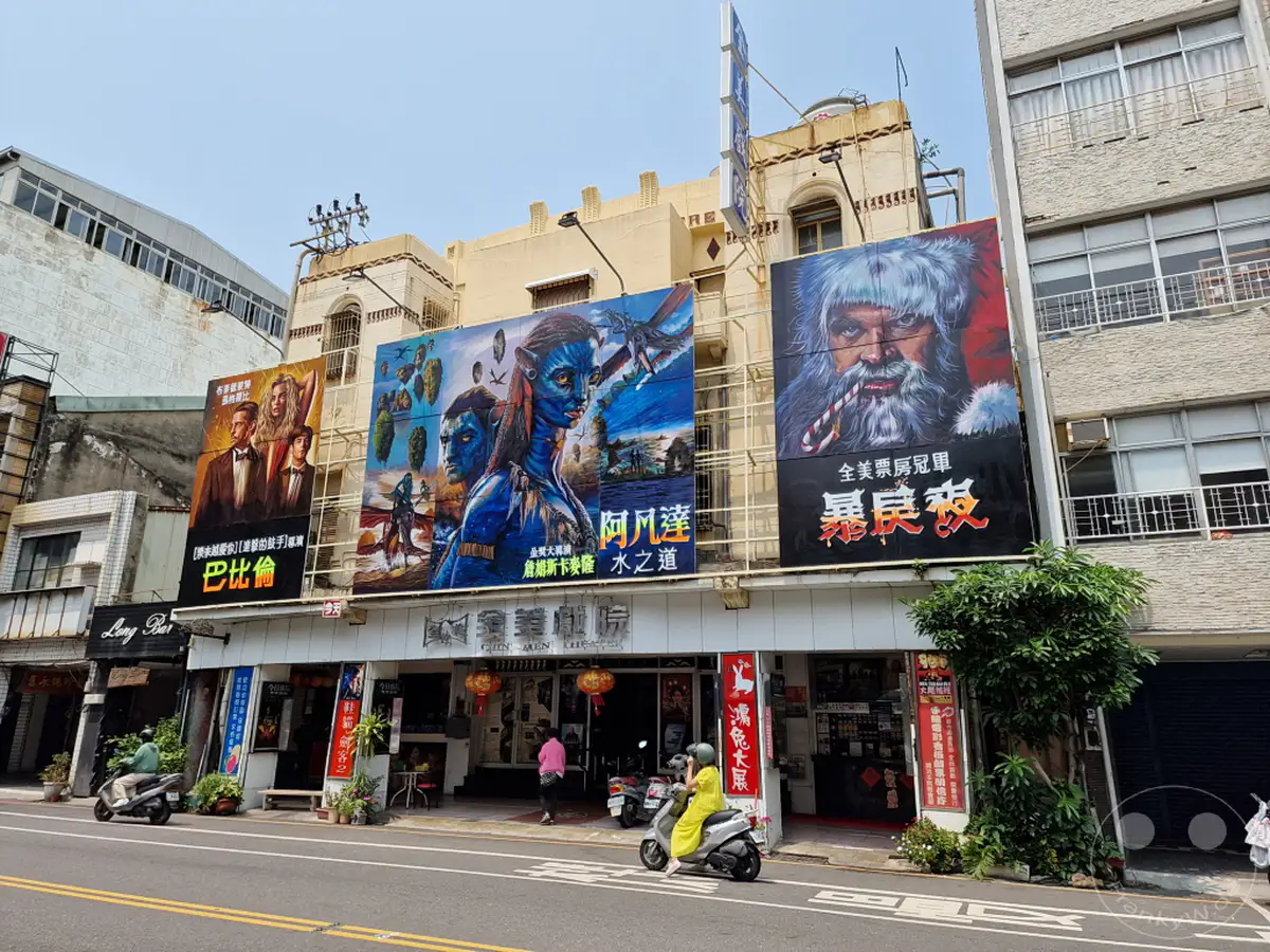 Vor dem Chin Men Theater in Tainan, Taiwan, werben großformatige, handgemalte Kinoplakate für aktuelle Filme und ziehen die Aufmerksamkeit von Passanten auf sich. Die Fassade des Theaters und die umliegende Bebauung prägen das urbane Stadtbild. Rollerfahrer bewegen sich auf der Straße vor dem Eingangsbereich, auf dem auch Lampions für zusätzliche Atmosphäre sorgen.