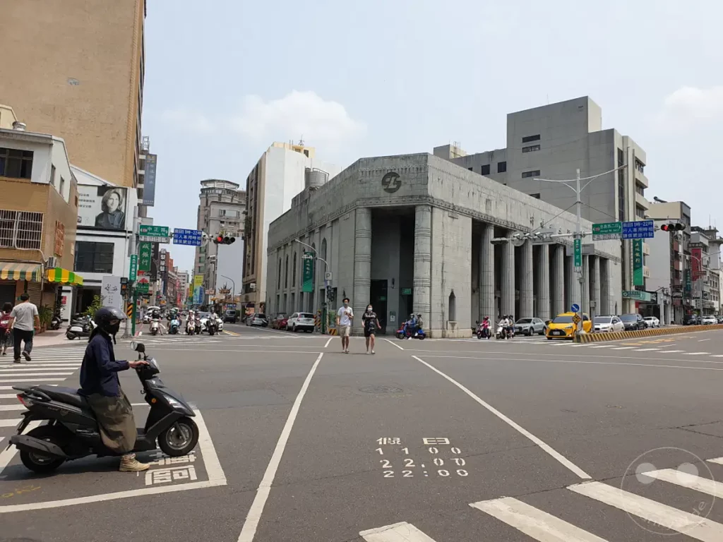 Ein belebter Straßenzug in Tainan, Taiwan, an einem sonnigen Tag. Autos und Motorroller bewegen sich durch eine Kreuzung, während Fußgänger auf den Zebrastreifen warten. Elegante historische Gebäude säumen die Straße und vermitteln ein Gefühl von urbanem Leben mit einem Hauch von Geschichte.