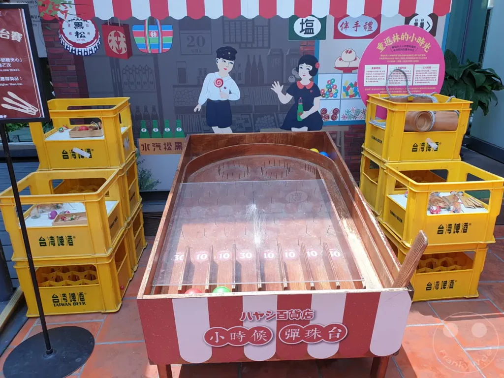 In Tainan, Taiwan, vor dem Hayashi Department Store steht ein nostalgisches Spielgerät, das an vergangene Zeiten erinnert. Bunte Bierkisten und eine liebevoll gestaltete Spielkulisse schaffen eine fröhliche und einladende Atmosphäre. Das Spiel, das an eine Art Murmelbahn erinnert, lädt zum Mitmachen und Verweilen ein und versprüht einen charmanten Retro-Flair.