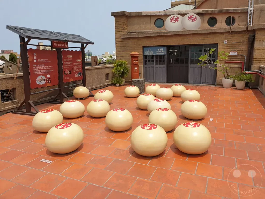 In Tainan, Taiwan, vor dem ehemaligen Hayashi Department Store, auch bekannt als "The Five Stories", stehen massive, kugelförmige Sitzgelegenheiten, die mit einem roten Muster verziert sind. Die orangefarbenen Ziegelsteine des Bodens und das helle Tageslicht schaffen eine sonnige und einladende Atmosphäre. Informationstafeln auf der linken Seite bieten Einblicke in die Geschichte des Ortes.