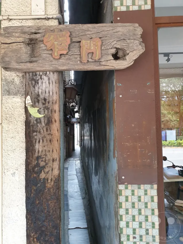 In Tainan, Taiwan, führt eine extrem schmale Gasse vorbei an verwitterten Holzelementen und alten Mauern. Die stimmungsvolle Szenerie strahlt eine Zeitlosigkeit aus und lädt dazu ein, verborgene Winkel der Stadt zu entdecken. Ein traditionelles Holzschild mit chinesischen Schriftzeichen markiert den Eingang zu diesem ungewöhnlichen Durchgang.