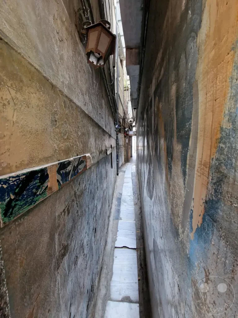 Eine extrem schmale Gasse in Tainan, Taiwan, flankiert von hohen Mauern, die mit verschiedenen Texturen und Graffiti verziert sind. Helle Steinplatten säumen den Weg durch die dunkle und atmosphärische Gasse. Eine alte Laterne hängt an der Wand und wirft Licht auf die ungewöhnliche urbane Szenerie.
