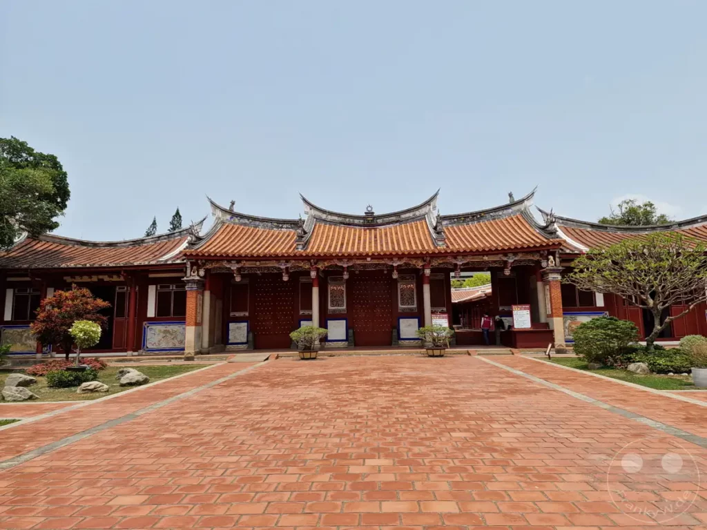 Ein traditioneller Konfuzius-Tempel in Tainan, Taiwan, präsentiert seine imposante Architektur mit geschwungenen Dächern und roten Holzsäulen. Der weitläufige, mit roten Ziegeln gepflasterte Vorplatz lädt zum Verweilen ein, während grüne Bäume und Pflanzen eine ruhige Atmosphäre schaffen. Die Szene vermittelt einen Eindruck von kultureller Tiefe und historischer Bedeutung.