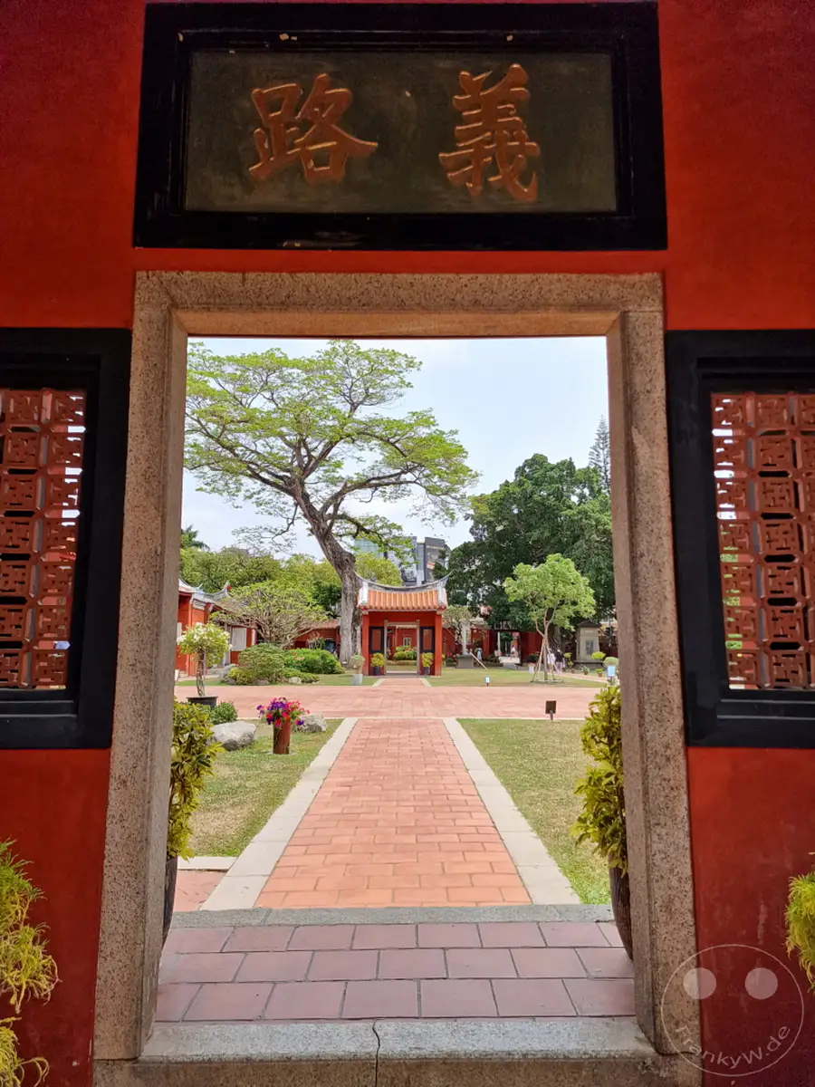 Ein Blick durch einen traditionellen Torbogen auf einen gepflasterten Weg, der zu einem historischen Konfuzius-Tempel in Tainan, Taiwan führt. Üppiges Grün und architektonische Elemente prägen die friedliche und kulturell reiche Szenerie. Die Atmosphäre ist ruhig und lädt zur Besinnung auf die reiche Geschichte und Tradition ein.