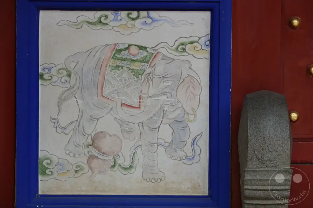 An der roten Holztür eines Tempels in Tainan, Taiwan, ist eine Malerei eines Elefanten eingerahmt von einem blauen Rand zu sehen. Die detailreiche Darstellung des Tieres mit seiner Decke und den Wolken im traditionellen Stil erzeugt eine feierliche Atmosphäre. Ein steinernes Postament steht rechts neben dem Eingang und vervollständigt die sakrale Kulisse.