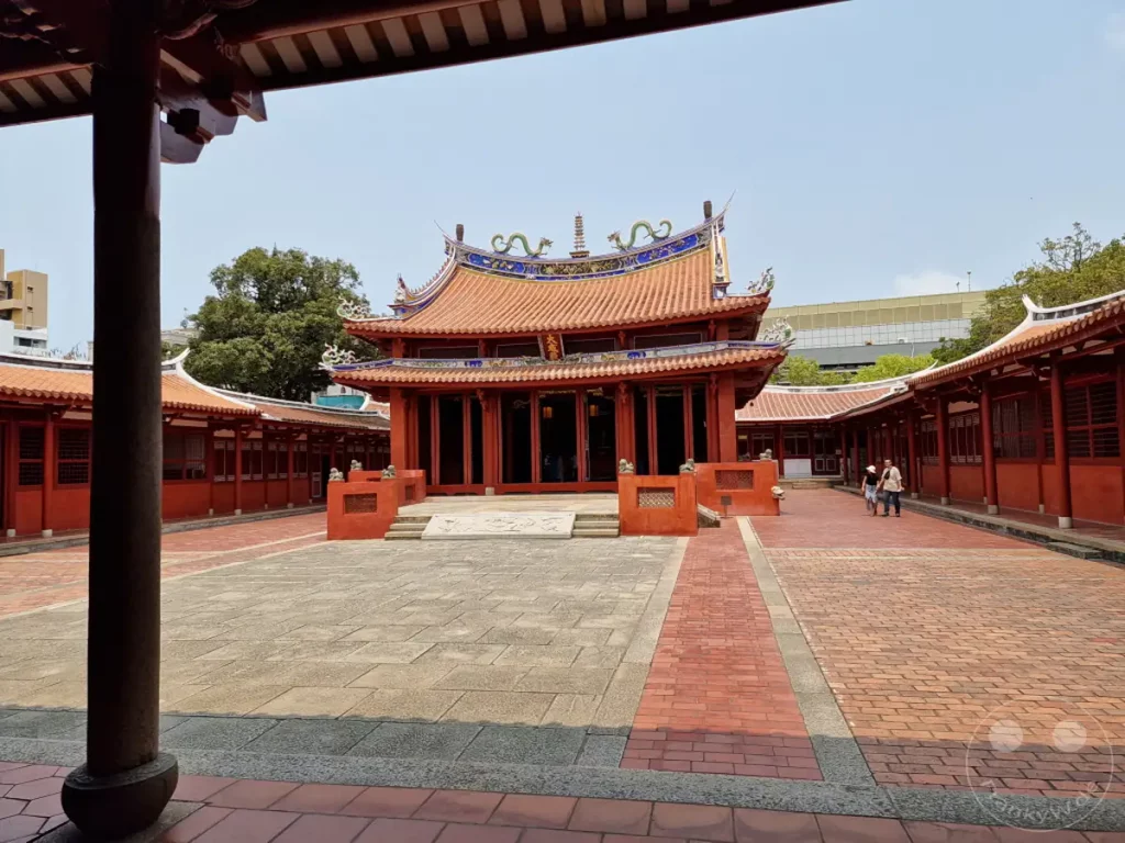 Im Hof eines traditionellen Tempels in Tainan, Taiwan, mit rotbraunen Säulengängen und einem zentralen Gebäude mit geschwungenen Dächern. Zwei Personen spazieren auf einem von Ziegeln gesäumten Weg durch die Anlage. Das friedliche Ambiente lädt zur Besinnung auf die Geschichte und Kultur ein.