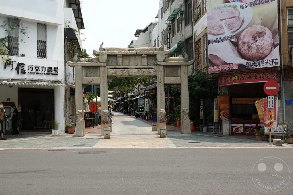 Ein traditioneller Torbogen aus Stein markiert den Eingang zu einer belebten Marktstraße in Tainan, Taiwan. Auf beiden Seiten säumen Geschäfte und Essensstände die Gasse, die von Einheimischen und Besuchern frequentiert wird. Die Szene strahlt eine lebendige Atmosphäre aus, die traditionelles und modernes Leben vereint.