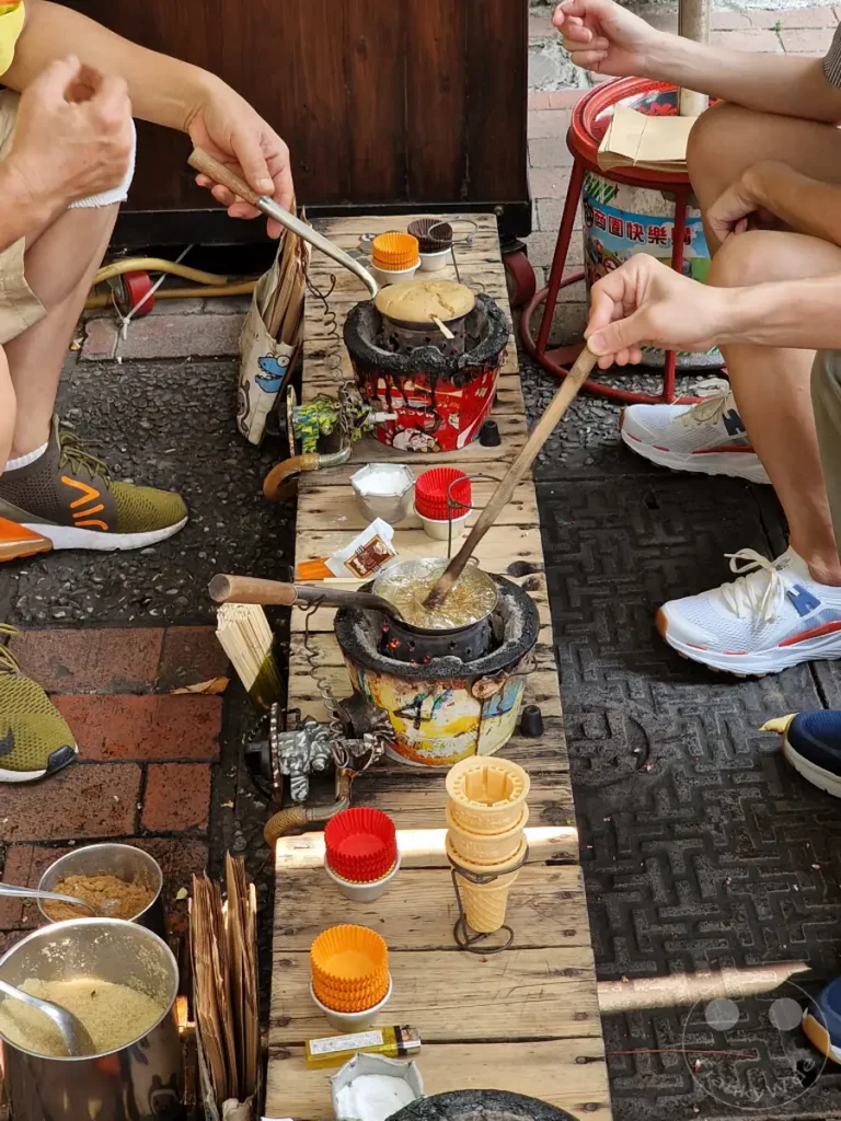 In Tainan, Taiwan, auf der Fuzhong Street, wird traditionelles Streetfood zubereitet. Hände bedienen kunstvolle Geräte, um süße Leckereien herzustellen, während die Atmosphäre geschäftig und authentisch ist. Kleine rote und orangefarbene Papierförmchen sowie Eiswaffeln warten auf die Fertigstellung der Speisen.