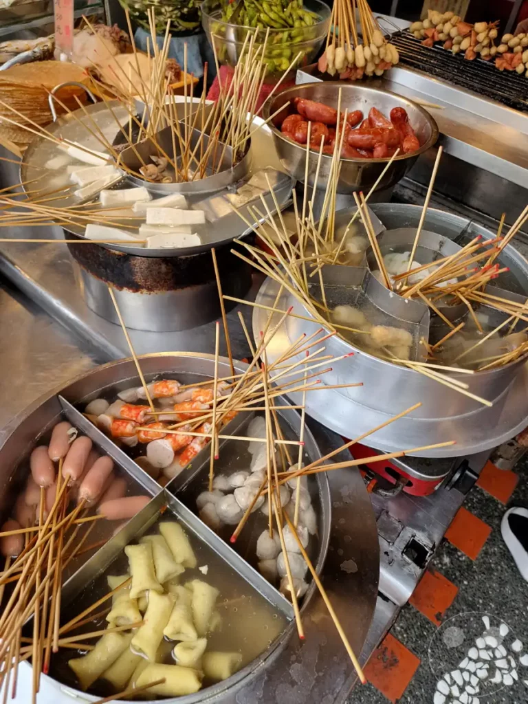 In Tainan, Taiwan, auf der Fuzhong Street, präsentieren sich verschiedene Garküchen mit einem vielfältigen Angebot an Streetfood. Die Bildkomposition fokussiert auf verschiedene Kochtöpfe, gefüllt mit dampfenden Leckereien wie Würstchen, Garnelen, Fischbällchen und Tofu, die an Holzspießen aufgereiht sind. Eine lebhafte und appetitanregende Szenerie, die typisch für die dynamische kulinarische Kultur Taiwans ist.