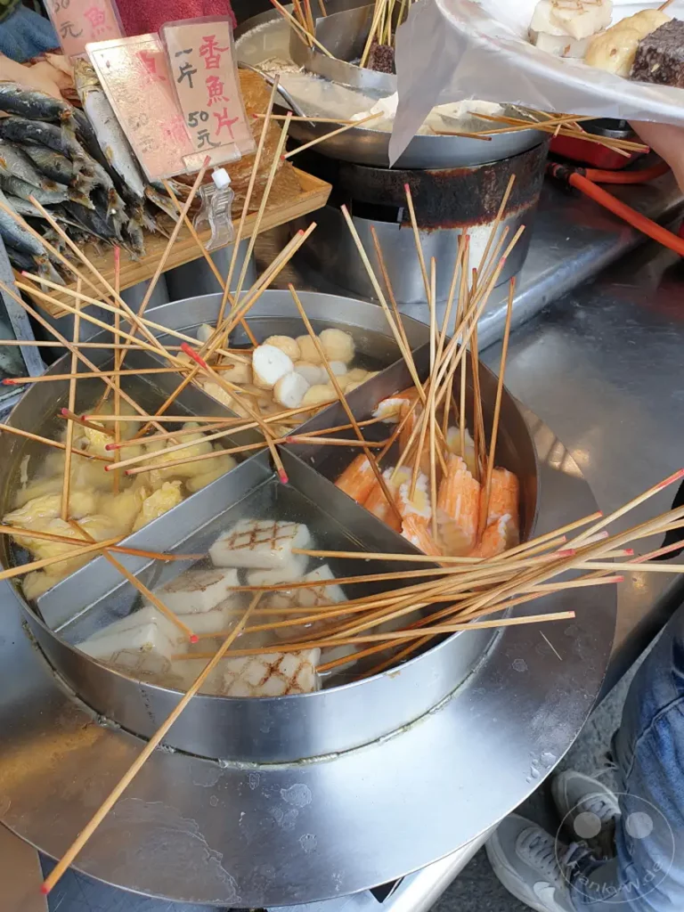 In Tainan, Taiwan, an der Fuzhong Street, werden auf einem Markt verschiedene Arten von Oden in einem großen Edelstahlbehälter angeboten. Zahlreiche Holzspieße ragen aus den dampfenden Portionen hervor, während im Hintergrund weitere Stände mit Fisch und anderen Speisen zu sehen sind. Eine authentische Streetfood-Atmosphäre wird durch die lebendige Darstellung der Speisenzubereitung vermittelt.