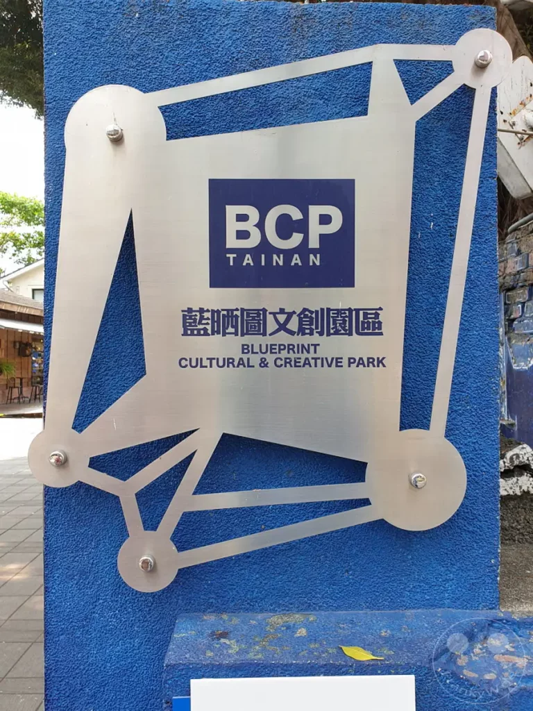Ein auffälliges Schild verkündet die Präsenz des Blueprint Culture & Creative Park in Tainan, Taiwan. Die metallische Struktur mit geometrischen Elementen ist vor einer lebendigen blauen Wand montiert und lädt Besucher ein, das kreative Umfeld zu erkunden. Die Szene strahlt eine Mischung aus moderner Kunst und kultureller Initiative aus.