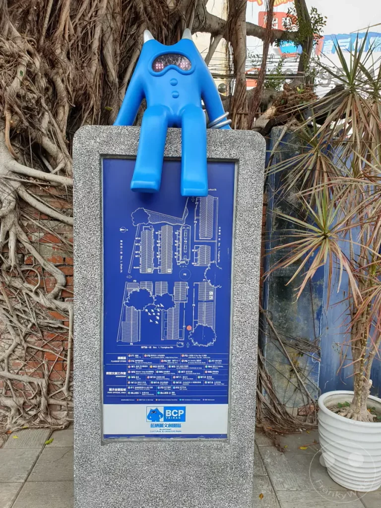 Eine blaue, menschenähnliche Skulptur sitzt auf einer Steinsäule mit einer blauen Karte oder einem Plan des Blueprint Culture & Creative Park in Tainan, Taiwan. Die Umgebung ist von den Wurzeln eines großen Baumes und tropischen Pflanzen umgeben und erzeugt eine atmosphärische, künstlerische Szene.