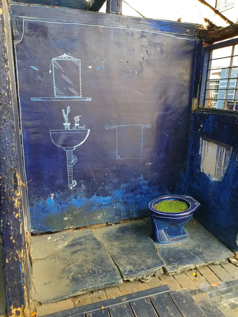 Im Blueprint Culture & Creative Park in Tainan, Taiwan, befindet sich eine provokante Installation, die eine Toilette als Pflanzgefäß nutzt und mit blau-weißen Linienzeichnungen Elemente eines Badezimmers andeutet. Die Szene wirkt trotz der kreativen Nutzung des Raumes leicht heruntergekommen und hinterlässt einen bleibenden Eindruck. Dieses Kunstwerk lädt zum Nachdenken über Natur und Urbanität ein.