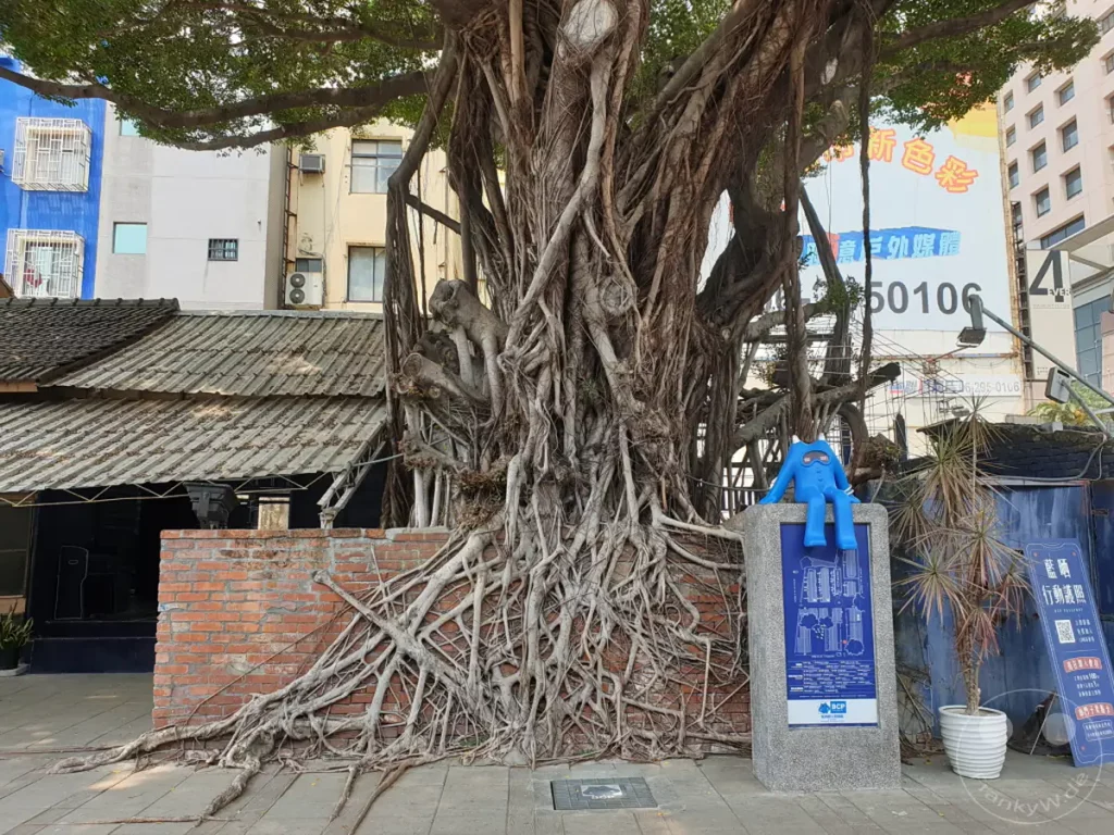 Ein imposanter Banyanbaum mit seinen charakteristischen Luftwurzeln dominiert die Szene vor einem historischen Gebäude in Tainan, Taiwan. Eine blaue, menschenähnliche Skulptur sitzt auf einem Sockel mit einer blauen Blueprint-Darstellung, die dem Ort seinen Namen gibt. Die Atmosphäre ist eine Mischung aus Naturwunder und künstlerischer Gestaltung, die zum Verweilen einlädt.