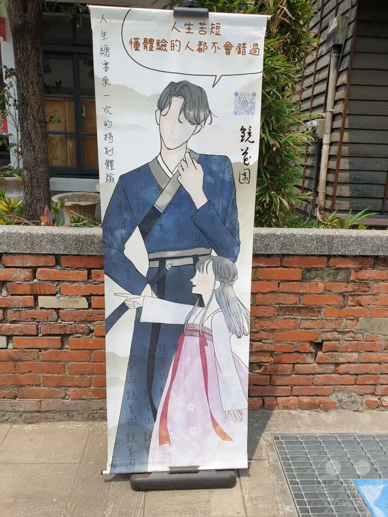 Ein Banner mit einer Illustration von zwei Personen in traditioneller Kleidung steht vor einer Ziegelmauer im Blueprint Culture & Creative Park in Tainan, Taiwan. Die Zeichnung zeigt eine aufwendig gestaltete Szene, die eine Geschichte erzählt und zur Reflexion über das Leben anregt. Die Umgebung, geprägt von rohen Ziegeln und üppigem Grün, schafft eine atmosphärische Kulisse für das Kunstwerk.