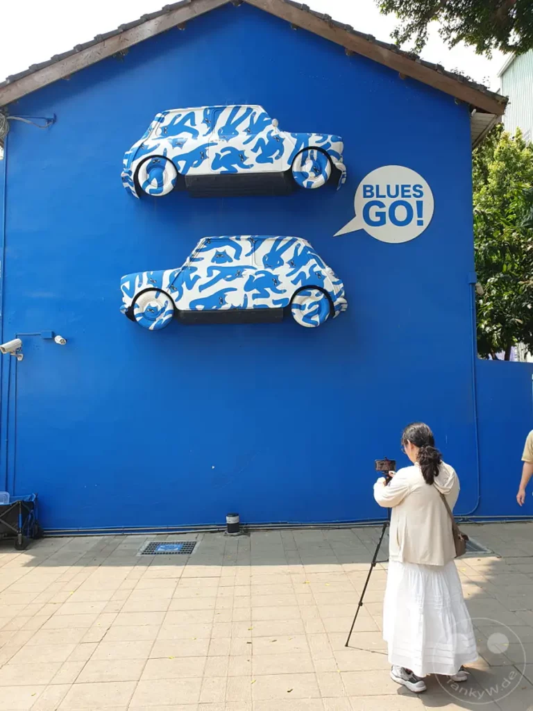 Zwei stilisierte Autos mit einem auffälligen blau-weißen Muster zieren eine knallblaue Wand auf dem Blueprint Culture & Creative Park in Tainan, Taiwan. Eine Frau filmt die Kunstinstallation mit ihrem Smartphone auf einem Stativ. Die Szene strahlt eine lebendige und kreative Atmosphäre aus, typisch für einen Kunstpark.