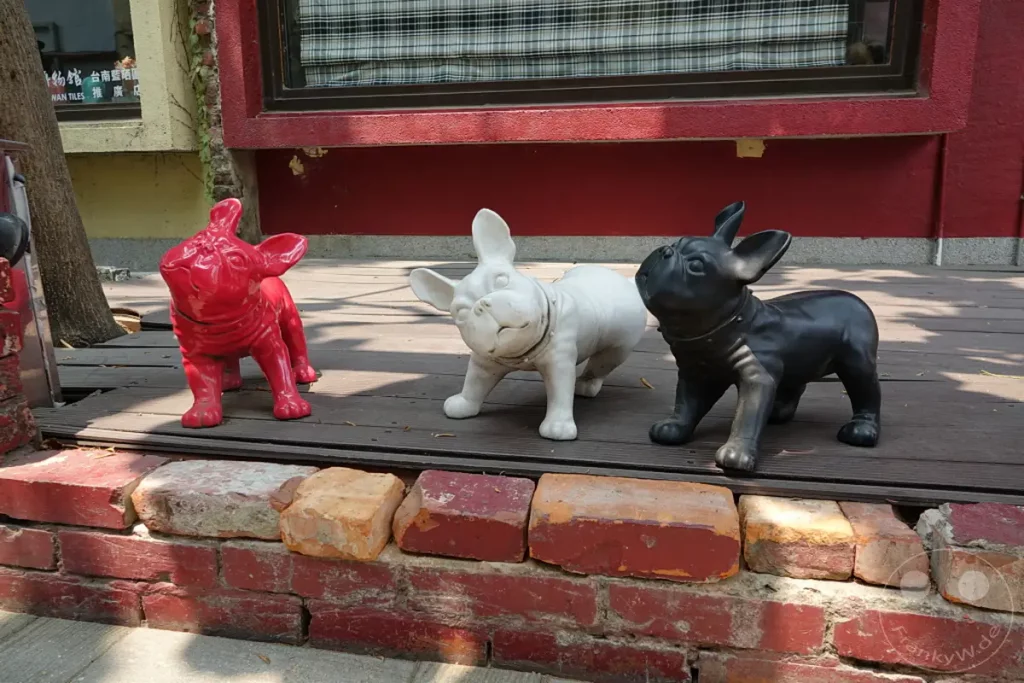 In Tainan, Taiwan, zieren drei charmante Französische Bulldoggen-Skulpturen den Außenbereich des Blueprint Culture & Creative Park. Die Dekofiguren in Rot, Weiß und Schwarz stehen auf einer Holzplattform, eingerahmt von einer Ziegelmauer, und verleihen dem Ort eine verspielte und künstlerische Atmosphäre.