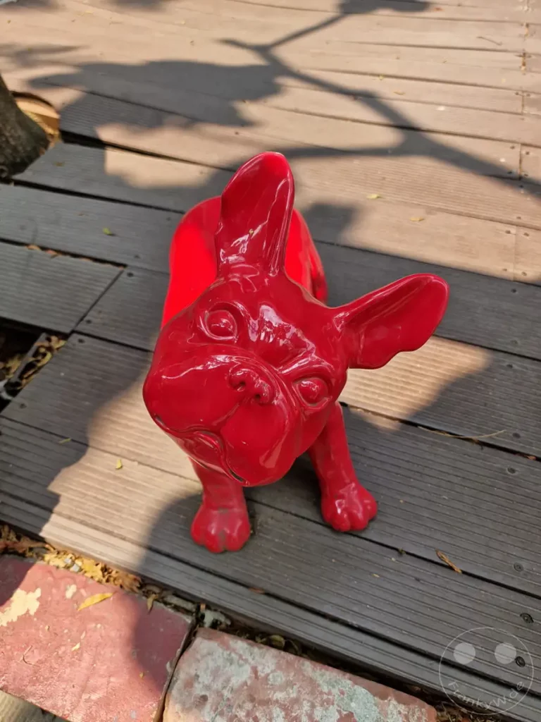 Eine glänzend rote Hundestatue, die an einen Mops oder eine Bulldogge erinnert, steht auf einem hölzernen Außendeck. Schatten von Ästen und Blättern fallen auf das Deck und die Skulptur und schaffen ein Muster aus Licht und Dunkelheit. Die lebendige Farbe der Statue hebt sich von den natürlichen Tönen des Holzes und der umgebenden Erde ab, was dem Ort eine verspielte und künstlerische Atmosphäre verleiht, die typisch für den Blueprint Culture & Creative Park in Tainan, Taiwan ist.