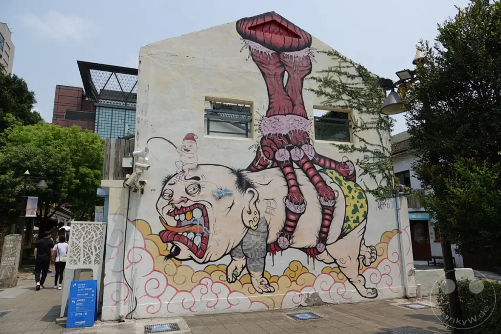Eine detailreiche Straßenkunst-Szene dominiert die Wand eines Gebäudes im Blueprint Culture & Creative Park in Tainan, Taiwan. Das bunte und surreale Wandgemälde stellt eine fantasievolle Kreatur dar, die auf einem grotesken Kopf reitet und eine düstere, aber faszinierende Atmosphäre schafft. Umgeben von grüner Vegetation und einem hellen Himmel fügt sich das Kunstwerk harmonisch in die urbane Umgebung ein.