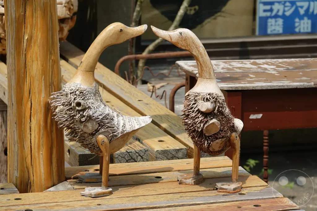 Zwei handgefertigte Entenfiguren aus Holz und Pflanzenfasern stehen auf einer Holzbank im Blueprint Culture & Creative Park in Tainan, Taiwan. Die Dekorationselemente scheinen sich einander zuzuneigen und erzeugen eine romantische und verspielte Atmosphäre. Die Szene fängt die charmante Handwerkskunst und die entspannte Stimmung des Parks ein.