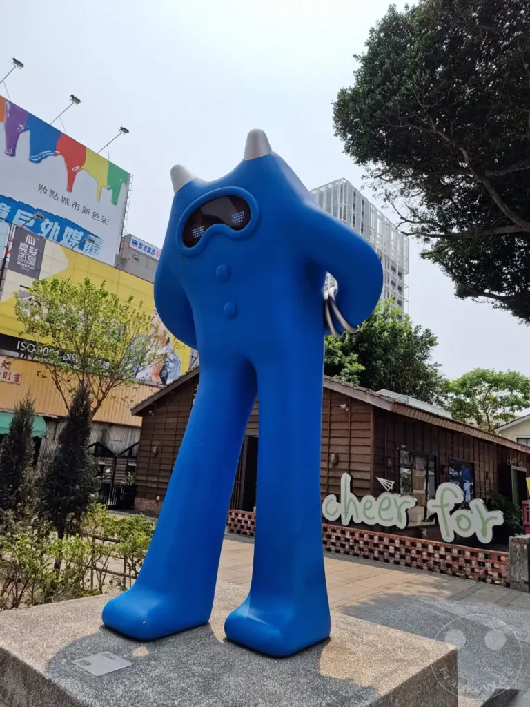 Eine auffällige, indigoblaue Skulptur dominiert die Szene im Blueprint Culture & Creative Park in Tainan, Taiwan. Hinter der Skulptur befindet sich ein charmantes Holzgebäude mit einer "cheer for"-Beschriftung, das von üppigen Bäumen und einer hellen, sonnigen Atmosphäre umgeben ist. Im Hintergrund sind eine hohe moderne Glasfassade und eine bunte Werbetafel zu sehen, die einen Kontrast zur rustikalen Ästhetik des Parks bilden.