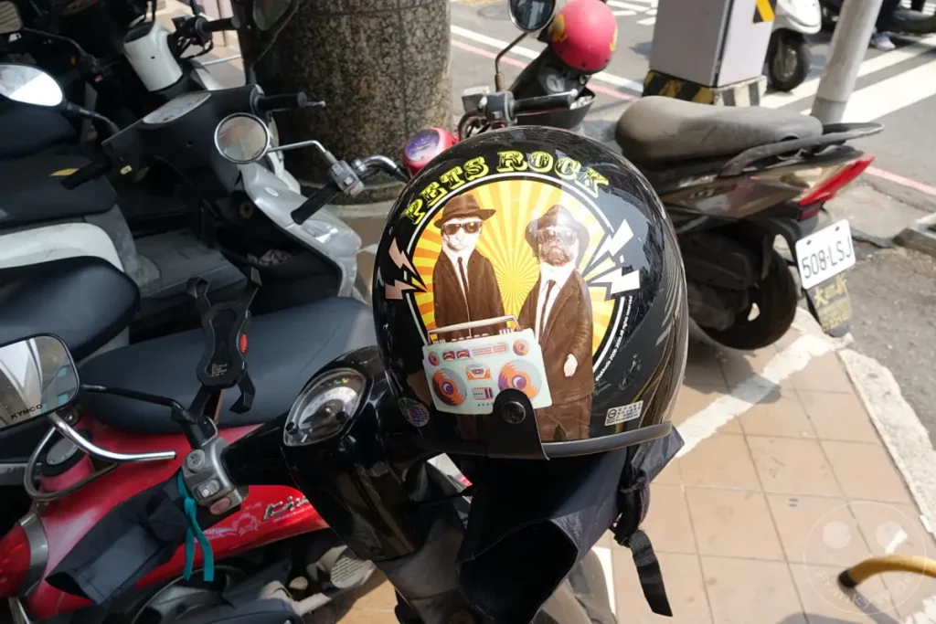 In Taiwan, Tainan, stehen mehrere Motorroller nebeneinander, einer davon ist mit einem auffälligen Helm verziert, der einen Kater und einen Mops mit Hüten und Sonnenbrillen zeigt. Der Helm wirbt für "Pets Rock" und trägt ein pastellfarbenes Boombox-Design, während im Hintergrund das architektonische Ambiente des Blueprint Culture & Creative Park zu erkennen ist. Die Szene vermittelt eine lebhafte und künstlerische Atmosphäre im Freien.