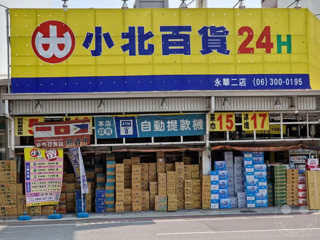 Ein bunter Supermarkt im Außenbereich in Tainan, Taiwan, mit einer gelben Fassade, die mit chinesischen Schriftzeichen und einem Logo verziert ist. Vor dem Geschäft sind zahlreiche gestapelte Getränkekisten und Werbebanner zu sehen, die eine geschäftige Atmosphäre vermitteln. Die Tageslichtaufnahmen und die gut sichtbaren Produkte deuten auf einen lebhaften Einzelhandelsort hin.