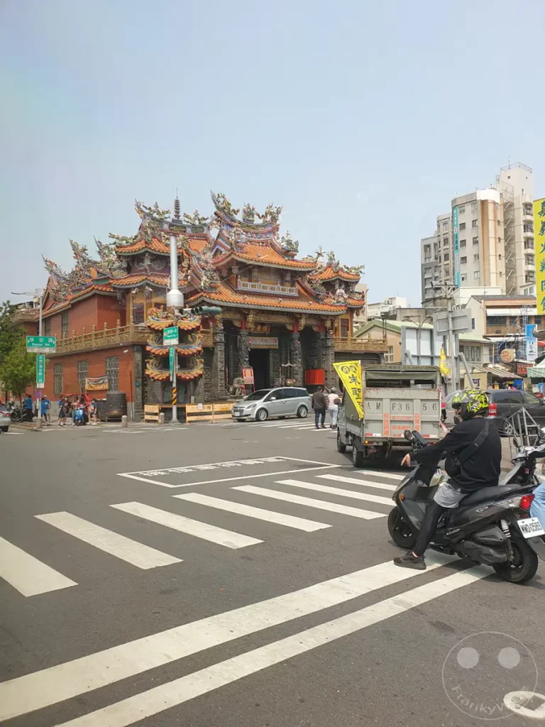 Eine belebte Kreuzung in Tainan, Taiwan, mit dem beeindruckenden South Plant Baoan Tempel im Hintergrund. Der Himmel ist klar und leicht bewölkt, während Fußgänger und Fahrzeuge den Kreuzungsbereich passieren. Die Szene vermittelt einen Eindruck des alltäglichen Lebens in dieser historischen Stadt.