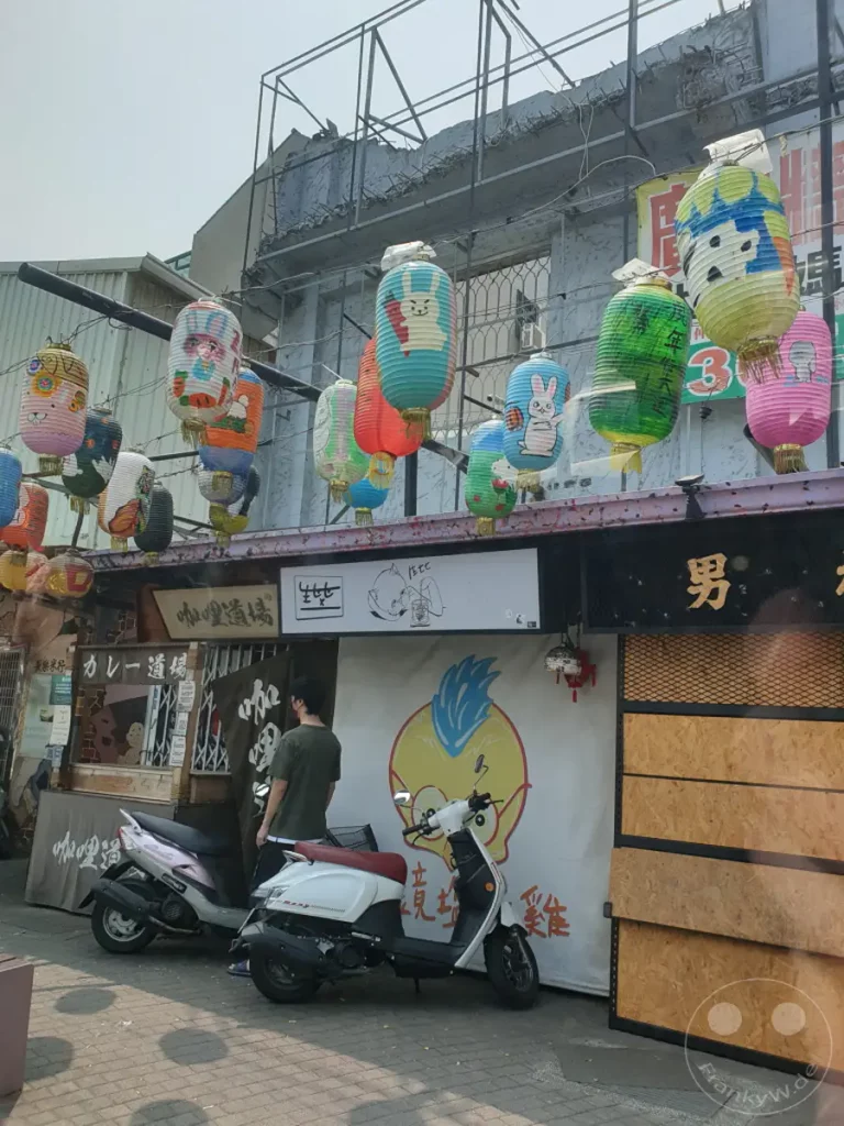Vor einer Hausfassade in Tainan, Taiwan, hängen bunte Papierlaternen mit Tiermotiven. Zwei Roller parken auf dem Gehweg vor den Geschäften, während eine Person mit dunklem T-Shirt im Hintergrund steht. Die Szene vermittelt eine alltägliche Atmosphäre in einer belebten Straße, akzentuiert durch die fröhlichen Dekorationen.