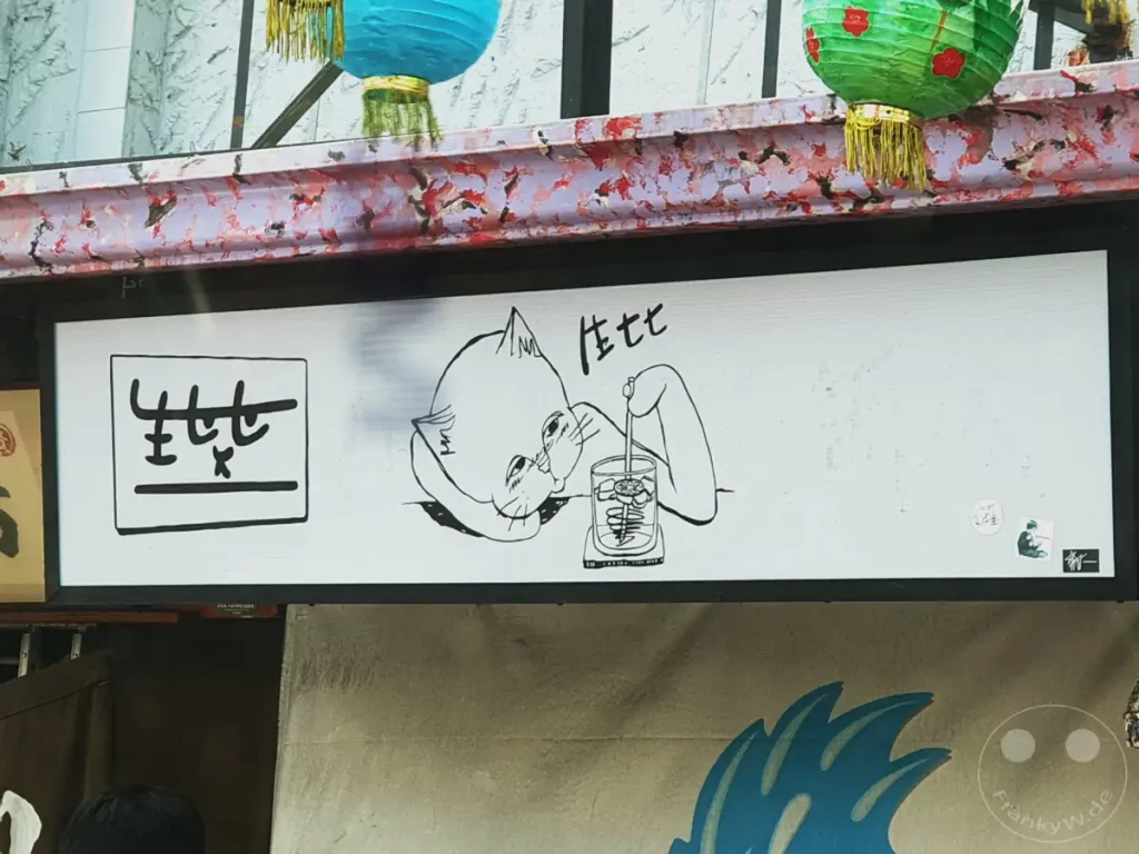 Ein Schild mit einer Zeichnung einer Katze, die mit einem Rührgerät in einem Becher hantiert, hängt über einem Geschäft in Tainan, Taiwan. Links daneben ist ein weiteres Symbol mit chinesischen Schriftzeichen abgebildet. Die Szene vermittelt eine humorvolle und künstlerische Atmosphäre.