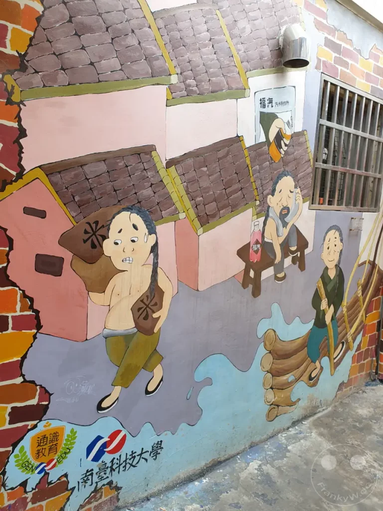 Ein buntes Wandgemälde in Taiwan, Tainan, stellt Szenen aus dem traditionellen Leben dar, mit detailreichen Darstellungen von Häusern und Figuren. Die lebendigen Farben und der satirische Stil erzeugen eine verspielte und nostalgische Atmosphäre. Die Kunstwerke auf der Mauer erzählen Geschichten aus einer vergangenen Zeit und beleben den städtischen Raum.