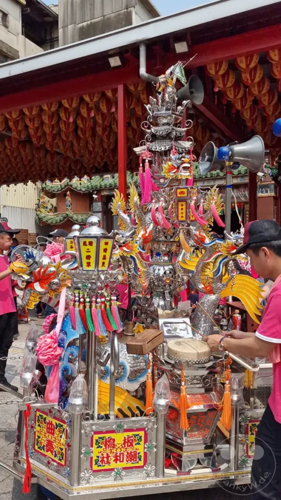 In Tainan, Taiwan, spielt ein Mann auf einer Trommel, während er Teil einer feierlichen Prozession ist. Ein aufwendig gestalteter Festwagen mit Drachenmotiven und bunten Quasten steht im Mittelpunkt der Szene. Die festliche Atmosphäre wird durch die Dekorationen und die Beteiligung der Menschen unterstrichen.