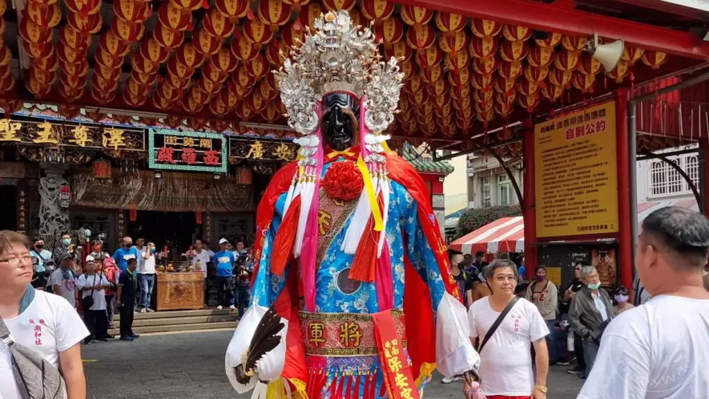 Eine farbenprächtige Prozession findet vor einem traditionellen Tempel in Tainan, Taiwan, statt. Eine aufwendig gestaltete Statue einer Gottheit wird im Mittelpunkt der Zeremonie getragen, umgeben von zahlreichen Gläubigen und geschmückt mit roten Laternen. Die Szene strahlt eine tiefe kulturelle Verbundenheit und spirituelle Atmosphäre aus.